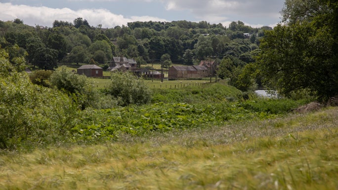 Several historic stone buildings of Kepier Hospital sit in the centre of the image, with trees in full leaf behind them, lush summer vegetation and a glimpse of the River Weir in the foreground. The scene is lit by sunlight under a blue sky with fluffy white and grey clouds.
